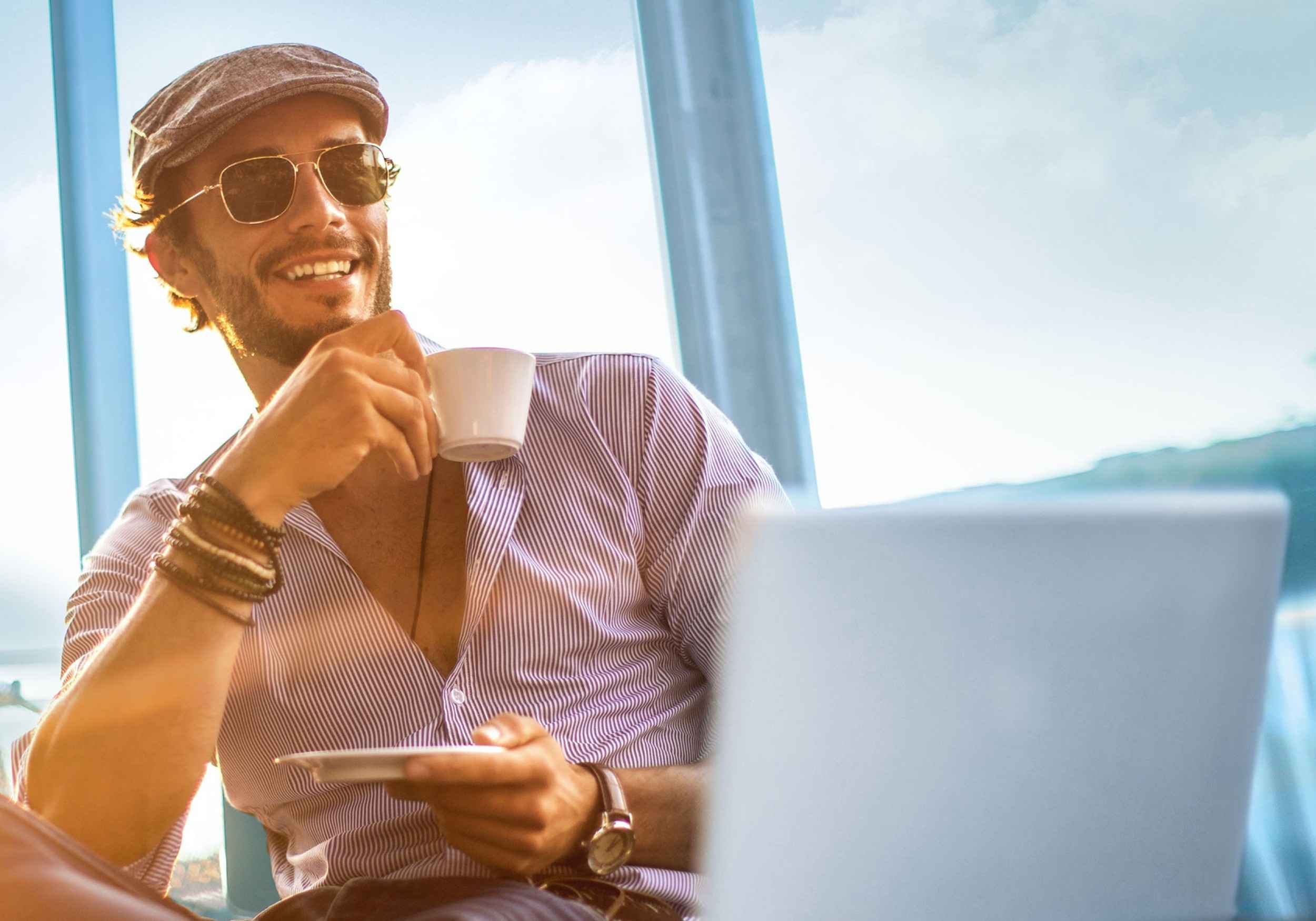 Mann mit Sonnenbrille und Kaffee in der Hand lächelt vor dem Laptop