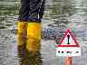 Ausschnitt von Beinen in Gummistiefeln neben Hochwasser-Schild im Hochwasser